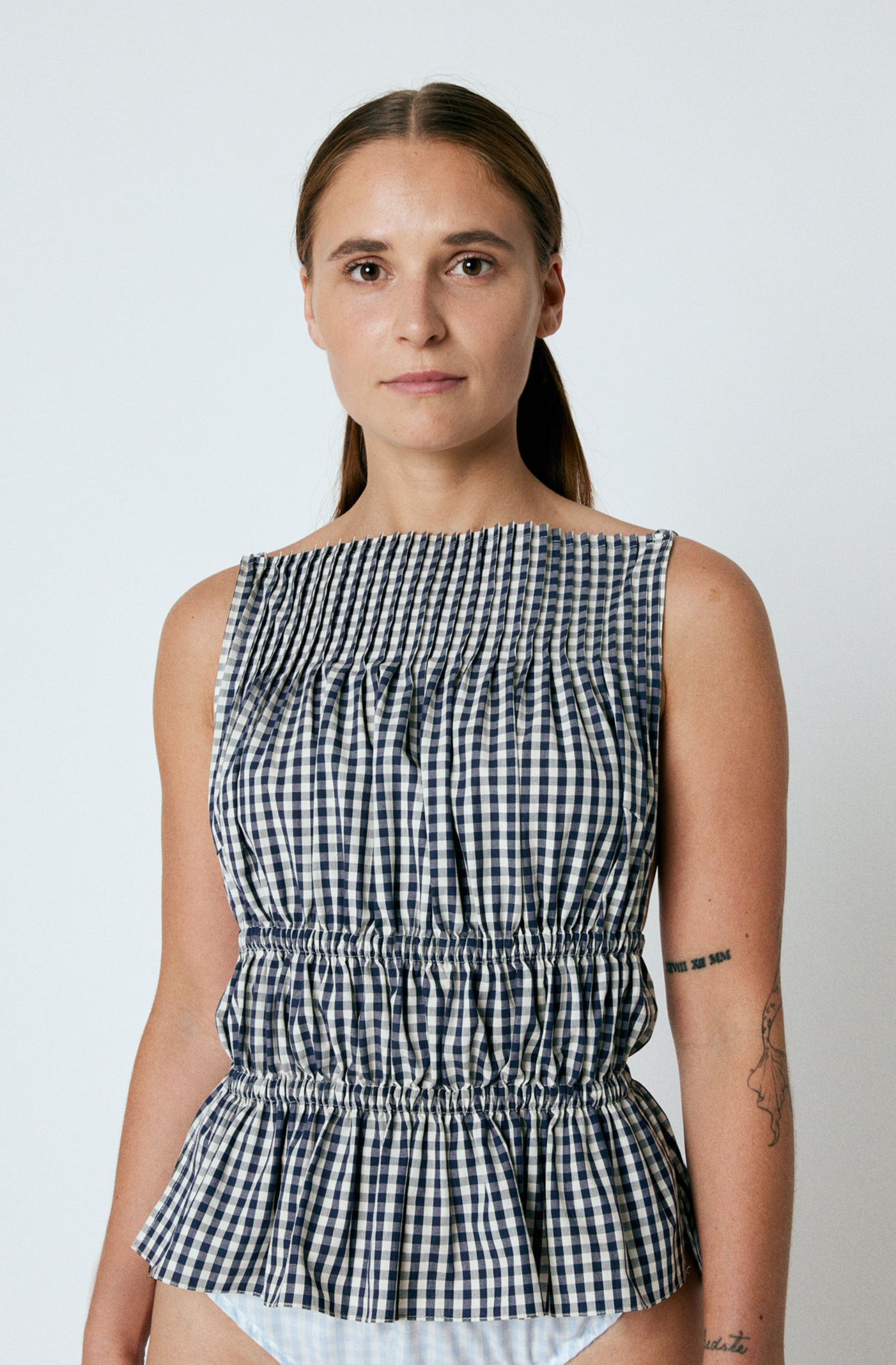 LINÉ Pintuck Top Navy. Pintucks at neckline · Elastic shoulder straps · Elastic casing under the bust and around the waist · Low back · The back hem is slightly elongated and is separated from the front by a small slit in the side seams · Model is 162 cm and wears size 1 · Made-to-order · Produced locally in Denmark · Material: 100 % organic cotton yarn dyed gingham · Collection: Gingham Garments. LINÉ