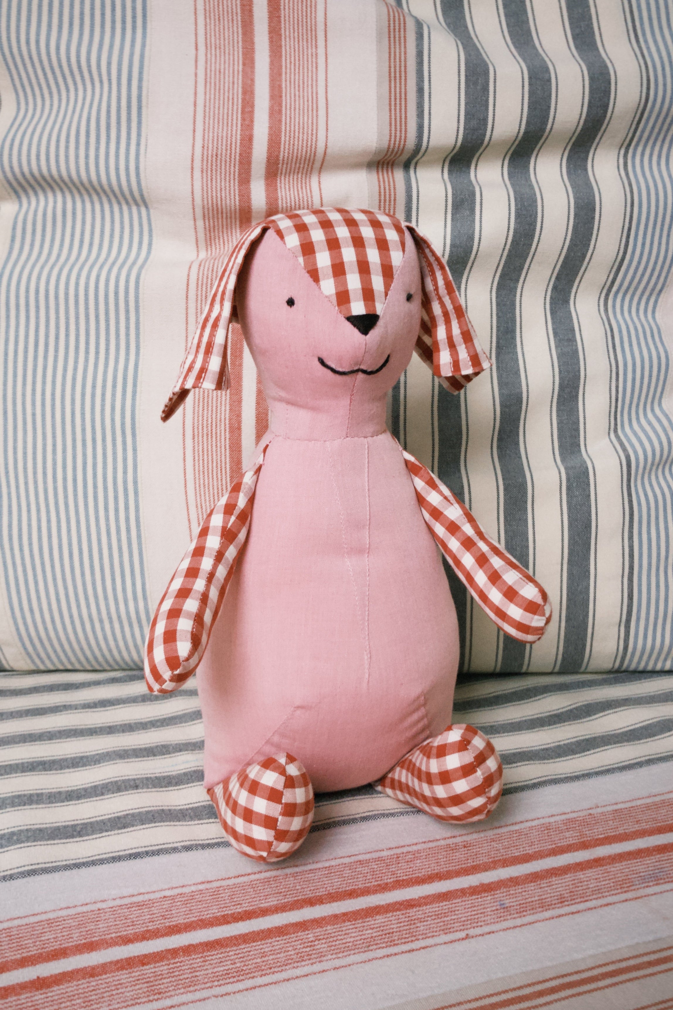 Pink teddy toy with checkered ears, arms and legs on a striped fabric background