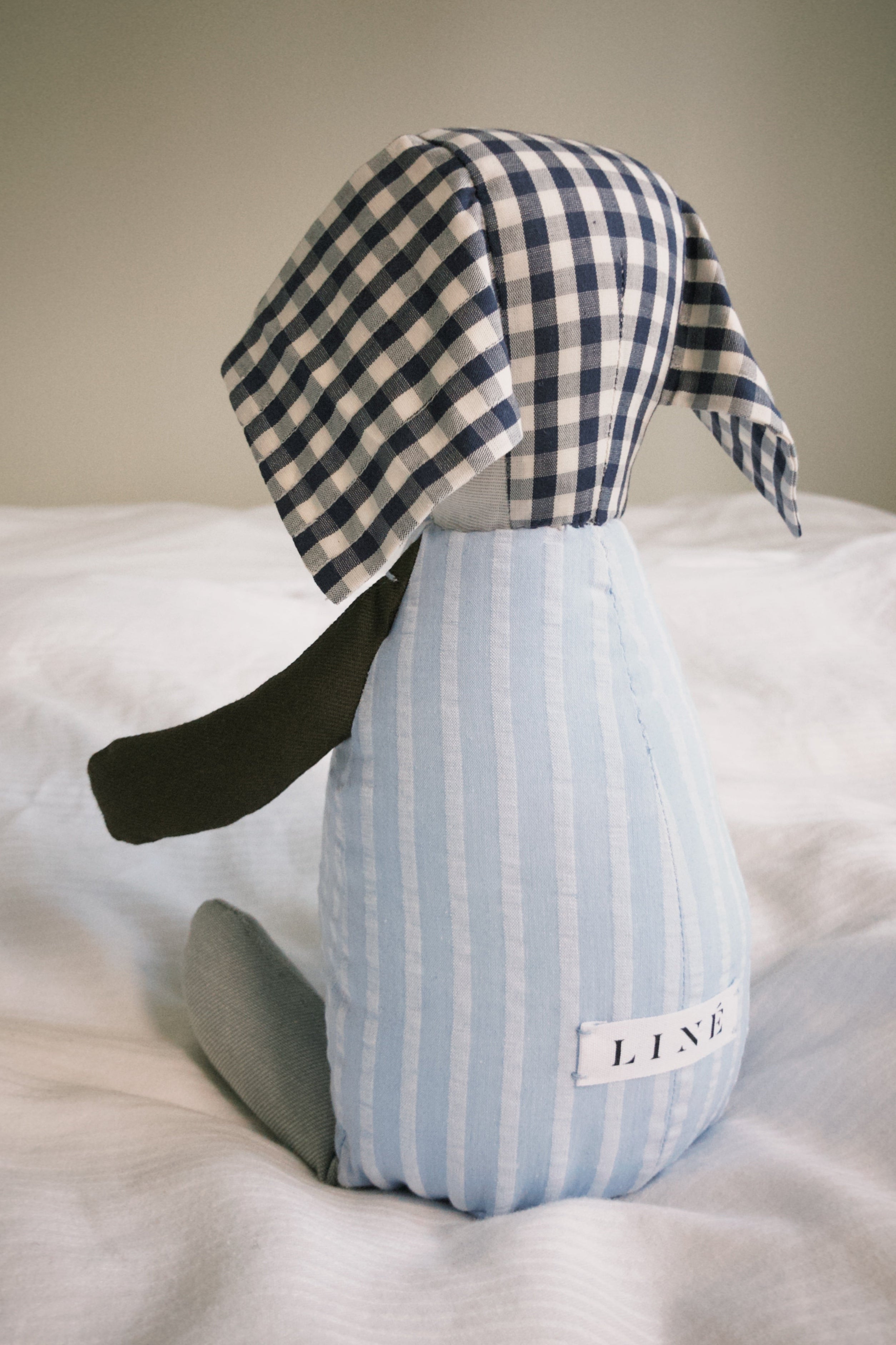 Teddy toy with checkered ears, striped blue body, brown arms and grey legs sitting on a bed