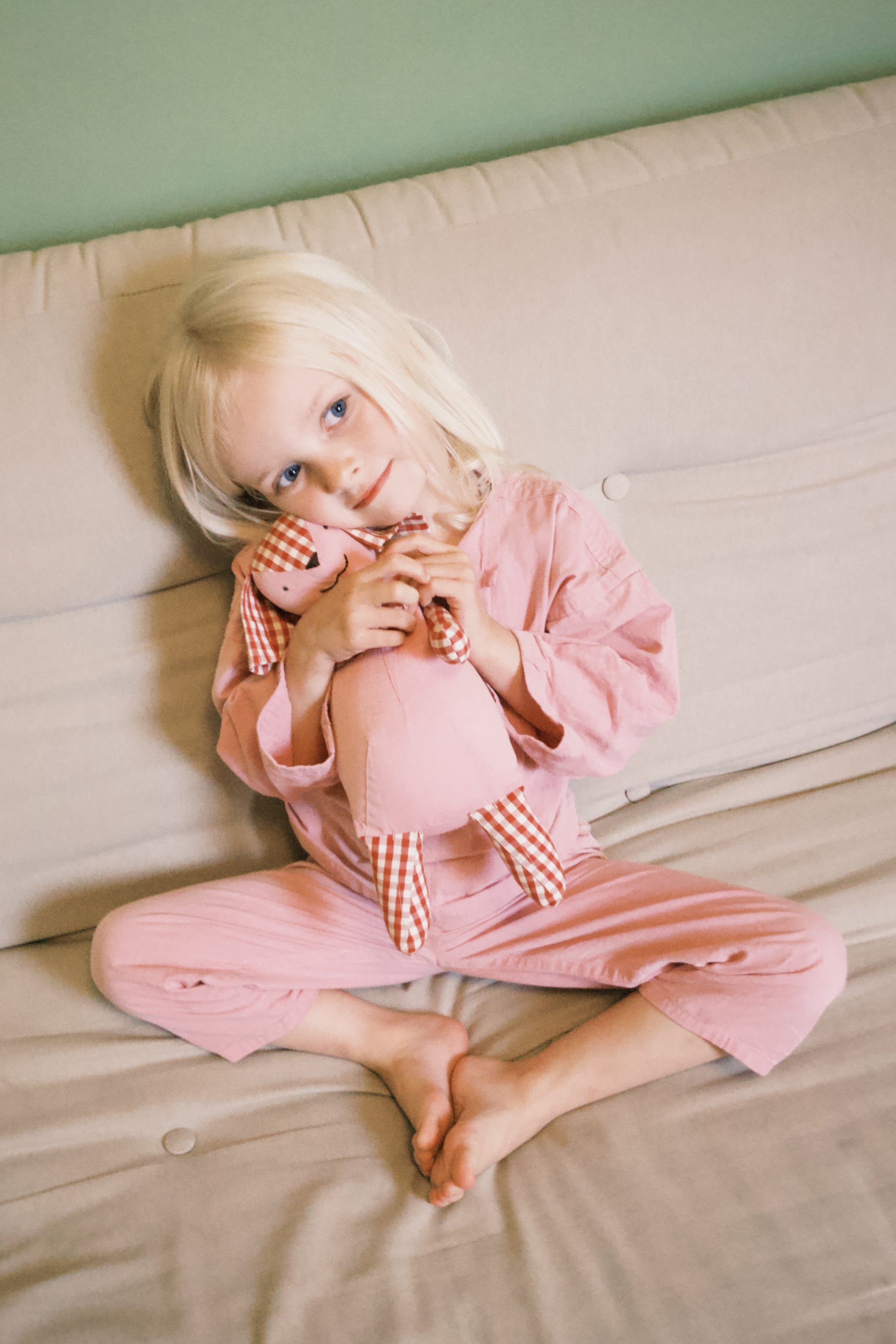 Child in pink pajamas sitting on a beige couch holding a ping and red checkered teddy toy.
